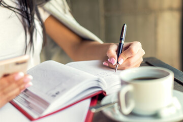 Business woman hand is writing in notepad with pen using smartphone in cafe.