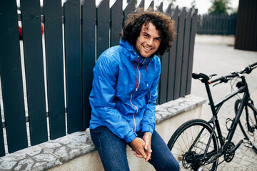 Fototapeta na wymiar Smiling Caucasian man with curly hair sitting on the fence with his bike before bicycling next to the house. Happy male courier with curly hair delivers parcel with a bicycle in the city.