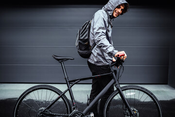 Outdoor side view image of a handsome man posing with his bike before bicycling on a rainy day next to the house. Happy male courier with curly hair delivers parcel cycling with a bicycle.