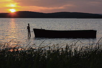 The silhouette of a wooden boat against the background of a Sunny sunset in summer, in the foreground reeds in the background a ridge of hills with the sun at the top.