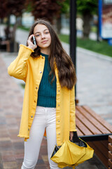 Fototapeta premium A young pretty teenage girl walks around the city in rainy weather. The girl is dressed in colored clothes and rubber boots talking on the phone