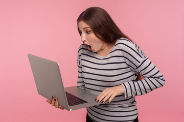 Portrait of surprised, frightened young woman in striped sweatshirt standing with open mouth, screaming in amazement and holding laptop, shocked by sudden info read. indoor studio shot, isolated