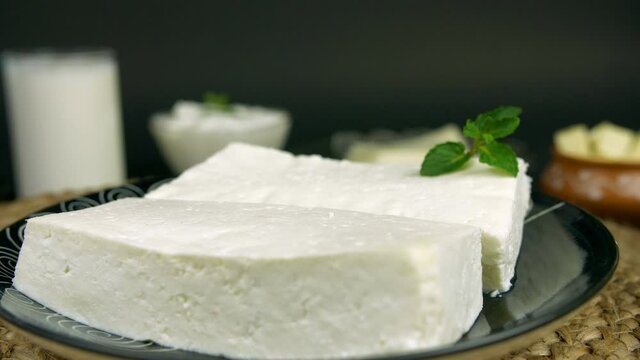 Closeup shot of fresh paneer rotating on a turntable in the clockwise direction. A black designer plate of white cheese slices placed on a kitchen mat against the greyish black background