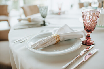 The table is covered with a white tablecloth, served. Plates, cutlery, napkin, pink glasses, seating cards, sprig of greens.