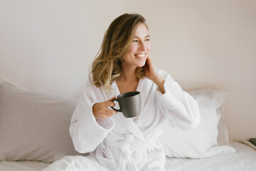Young beautiful woman wearing white bathrobe having breakfast in bed with coffee and croissant and...