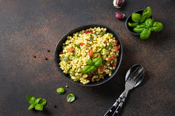 Vegetable salad with pasta Ptitim or Birdy, Israeli couscous decorated basil leaves on dark. Top view.