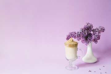 Beautiful dalgona drink a frothy coffee in a transparent mug and flowers lilac to purple Il purple background. Next to the mug is a branch of lilac flowers in a white vase. Copy space