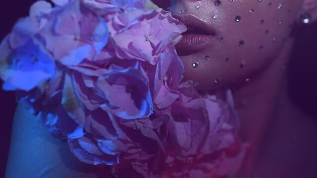 Beauty fashion Woman face decorated with gem stones, crystals. Closeup Portrait with Hydrangea, neon, UV lights. Model girl, holiday Glamour shiny make up with gems, jewellery, jewelry, accessories