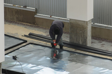 The men's hands are in gloves, twisting the cutter into a sheet of iron with an electric screwdriver. Outdoor. Without the involvement of workers, repairs are done independently