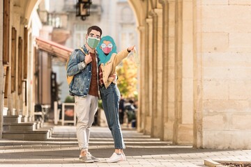 Obraz na płótnie Canvas couple of tourists with illustrated faces in medical masks hugging on street and pointing with finger away