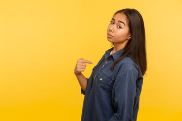 This is me! Portrait of arrogant selfish girl in denim shirt proudly pointing herself, boasting of success, feeling supercilious and self-confident. indoor studio shot isolated on yellow background