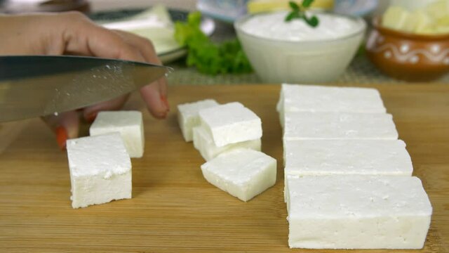 Closeup shot of woman hands cutting fresh paneer on a wooden chopping board. Side view of an Indian female slicing white cheese/paneer using a knife on the kitchen counter - dairy product