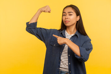 Look, I am strong! Portrait of confident proud girl in denim shirt pointing at biceps, expressing...