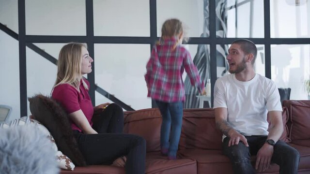 Joyful little girl running to parents sitting on couch and jumping on sofa. Cheerful Caucasian child having fun on weekends with young father and mother. Joy, leisure, unity, lifestyle, love.