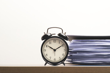 Black vintage alarm clock on working desk with many paper files stacked in black folder isolated on white wall background.