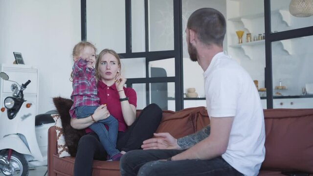 Young woman and little girl scolding brunette man with finger. Portrait of mother and daughter gesturing and joking. Happy joyful Caucasian family sitting on couch at home. Unity, fun, leisure.