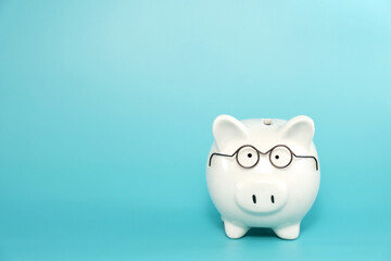 White ceramic piggy bank wearing reading glasses on blue teal background. Concept for money savings plan for retirement, aged society, or financial accounting