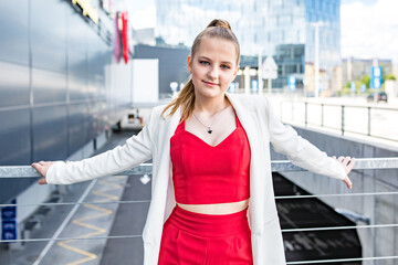 young blonde schoolgirl in a red suit and a white jacket posing in the parking lot