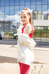 young blonde schoolgirl in a red suit and a white jacket posing in the parking lot