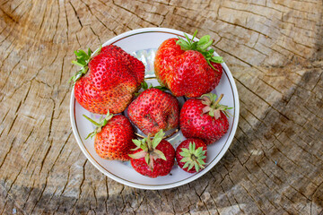 Red strawberries on a beautiful white porcelain plate. Rustic big and small, delicious and flavorful. With green stalks. On a wooden saw.