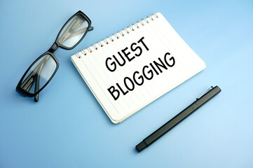 glasses,pen and notebook written guest blogging over blue background. Concept meaning used by the owners of blogs to increase traffic to their site.