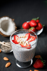 Chia seeds pudding with strawberries and coconut chips in glass on dark background.