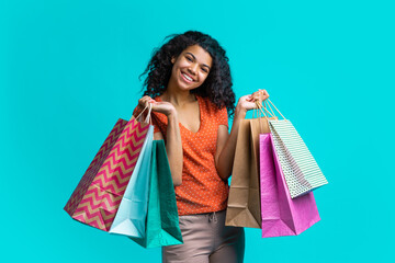 Cute dark skinned girl with perfect smile posing with piles of shopping bags isolated over bright blue background