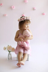 A cute girl in a wreath hugs a Teddy bear. The baby is sitting on a high chair and looks away.