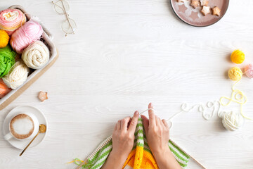 Hands of craftswoman knit a bright knitted striped fabric. Woolen and cotton yarn lies in a basket. Cappuccino coffee, cookies and glasses lie on a white wooden background.