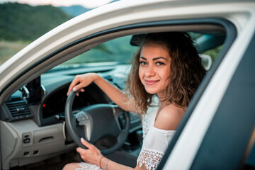 Obraz na płótnie Canvas Smilling young girl in white dress driving a car. Attractive girl sits in the driver's seat in automobile, outdoors summer portrait. Caucasian girl.