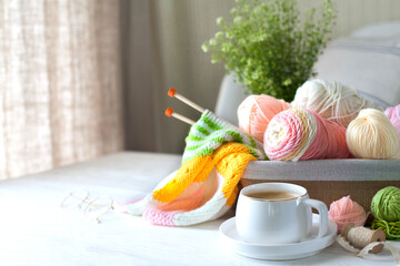 Knitted bright striped fabric. Woolen and cotton yarn lies in a basket. Cappuccino coffee, cookies and glasses lie on a white wooden background.
