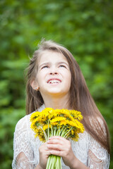 A fashionable girl in a beautiful white dress poses with yellow dandelion flowers in green foliage
