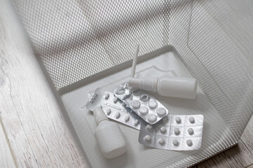 Medication in a white mesh metal wastebasket. Pills thrown into the trash. A paper basket with medicines inside on a wooden floor.
