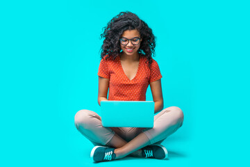 Beautiful young smiling woman in casual outfit and trendy eyeglasses sitting isolated on bright colored blue background and working on her laptop - obrazy, fototapety, plakaty