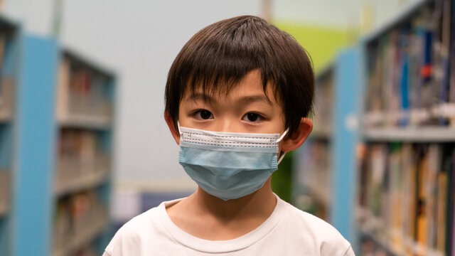 One cute Asian boy is wearing a disposal surgical mask at the public library. Kid and virus protection at the school.