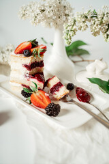 Sliced white berry cream cake decorated with strawberries and blackberries, among lilac flowers and green leaves. Food photography. Advertising and commercial close up design.
