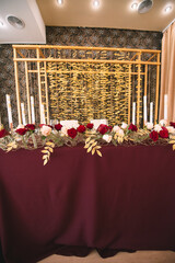 Presidium, banquet table with marsala tablecloth, candles, setting cards, napkins, plates, cutlery, glasses, composition of flowers of marsala rose. Candles, decor in gold, wine and marsala colors