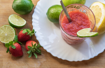 A glass of red cocktail with fruit on the white plate and wooden table. Apple, lime, lemon, strawberry. Spoon inside the glass.