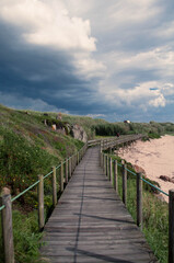 Fototapeta na wymiar Wooden walkway at the seaside under dark clouds, before thunderstorm sky. High grass along the shore
