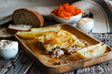 Tasty casserole with meat, potatoes, cheese and spices. Casserole served with carrot, bread and sour cream. Russian traditional dish. Dish on a wooden table