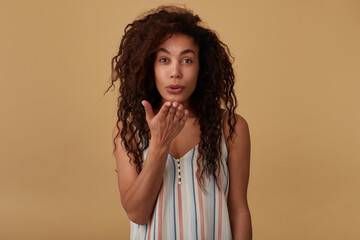 Indoor shot of positive young lovely dark skinned brunette woman with loose hair forming her lips in kiss and keeping palm raised while standing over beige background