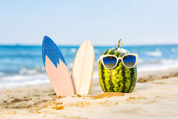 Ripe attractive watermelon surfer in stylish mirrored sunglasses with surfboard on sand against...