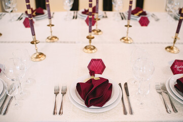 Banquet table with white tablecloth, candles, setting cards, napkins, plates, cutlery, glasses, composition of flowers of marsala rose. Candles, decor in gold, wine and marsala colors