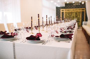 Banquet table with white tablecloth, candles, setting cards, napkins, plates, cutlery, glasses, composition of flowers of marsala rose. Candles, decor in gold, wine and marsala colors