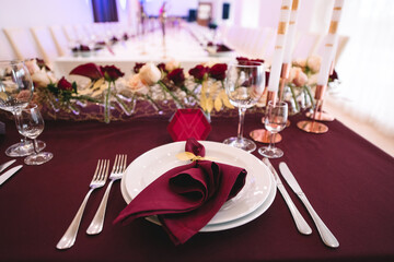 Banquet table with marsala tablecloth, candles, setting cards, napkins, plates, cutlery, glasses, composition of flowers of marsala rose. Candles, decor in gold, wine and marsala colors