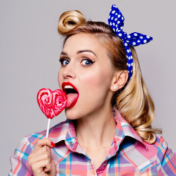 Portrait of woman licking heart shape lollipop dressed in pinup style, over grey background. Caucasian blond girl in retro fashion and vintage concept. Square composition.