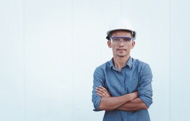 Thai male engineer wearing helmet and glasses standing with arms crossed In front of the container.
