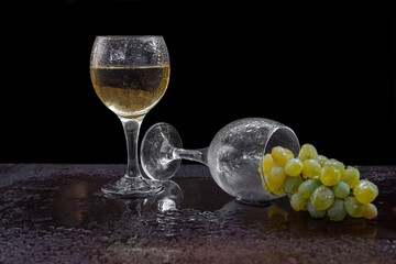 Two glasses of wine and an empty one lies on the table with a bunch of white grapes and drops of water on a black background