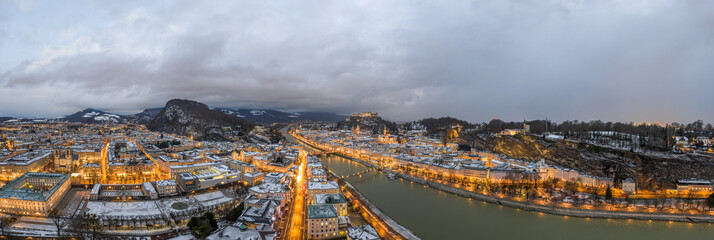 Panoramic aerial drone view of Salzburg snowy old town with city light on at dusk hour in winter