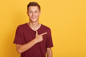 Young student male pointing aside with his index finger, wearing casually, looking smiling at camera, standing isolated over yellow background. Copy space for advertisement.